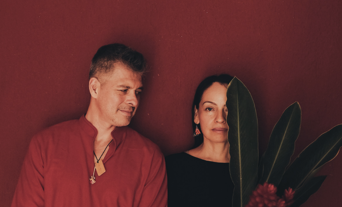 Mexican acoustic guitar duo Rodrigo y Gabriela posing against a deep red wall with tropical plant leaves in a promo photo.